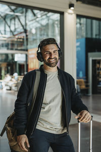 Portrait of happy man wearing bluetooth headphones while standing with luggage at station