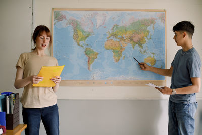 Teenage girl reading while boy pointing on world map in classroom