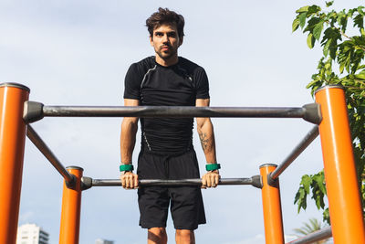 From below focused hispanic male athlete exercising on monkey bars against lush trees during fitness workout on sports ground in park