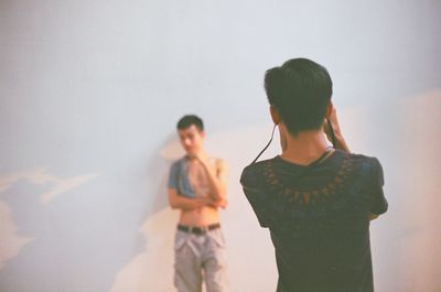 Rear view of man photographing friend standing against wall
