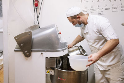 Male chef holding bowl while using electric mixer in bakery kitchen during covid-19