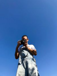 Low angle view of man photographing through camera while standing against clear blue sky
