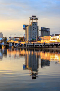 Illuminated factory by river against sky during sunset