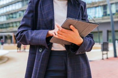 Low angle cropped unrecognizable female in stylish formal suit browsing digital tablet while standing against contemporary office building in downtown