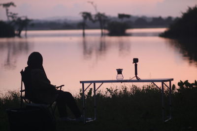 Rear view of silhouette man sitting by lake against sky during sunset