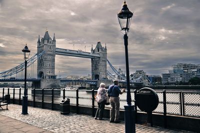 Full length rear view of senior couple on sidewalk by tower bridge against cloudy sky