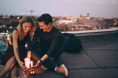 Smiling young couple protecting burning candle with hands while sitting on terrace against sky during dusk