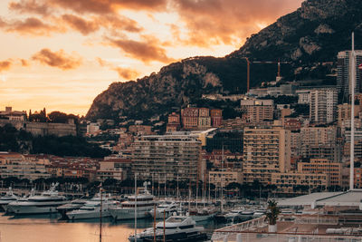 Sailboats moored at harbor by buildings against sky during sunset