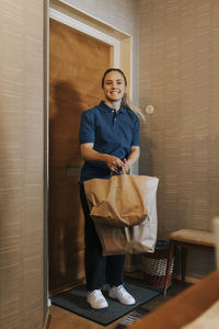 Smiling female care assistant holding bags while standing near door at home