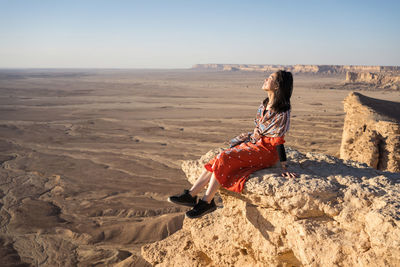 Side view of ethnic asian woman in casual clothes sitting on cliff with closed eyes contemplating the views in the edge of the world in saudi arabia