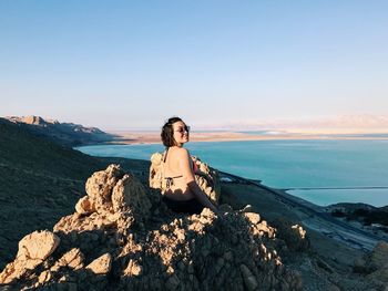 Rear view of woman wearing bikini while sitting on mountain by sea against sky