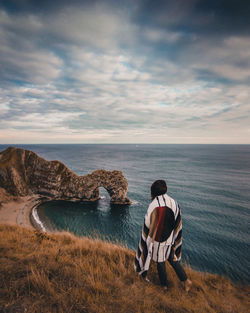 Rear view of man sitting on rock by sea against sky