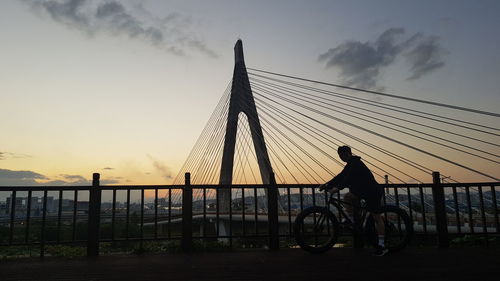 Silhouette man riding bicycle on bridge against sky during sunset