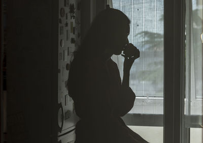 Silhouette woman looking through window at home