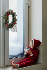 A thoughtful boy in a santa costume sits at the window and makes christmas decorations