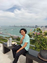 A young man sits with two thumbs up with the atmosphere behind the city and the sea.