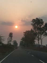 Road by trees against sky during sunset