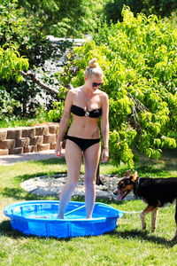 Sensuous woman in bikini standing in wading pool by dog in back yard