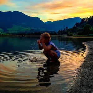 Boy washing face in lake against cloudy sky during sunset