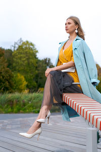 Happy woman swinging on swing in city park, wearing yellow dress and blue coat