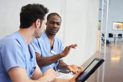 Male nurses discussing over digital tablet while sitting in corridor at hospital