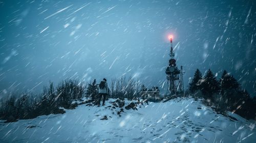 Rear view of man walking on snow covered land by illuminated communication tower against sky