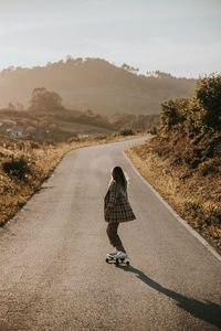 Back view faceless sporty female in trendy wear riding cruiser board along empty asphalt road in summer countryside on sunny day