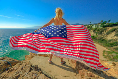Rear view of mature woman holding american flag while standing on cliff by sea against blue sky