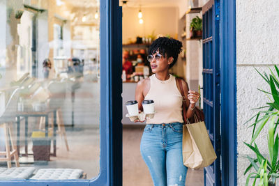 Positive young african american female with curly hair in casual clothes while leaving modern cafe with takeaway coffee cups and paper bags