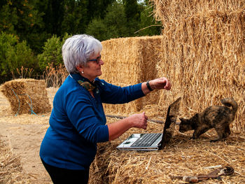 Woman playing with cat by laptop on hay bales 