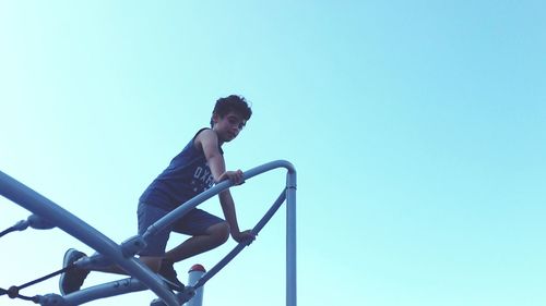 Low angle view of boy climbing on outdoor play equipment against clear blue sky at playground