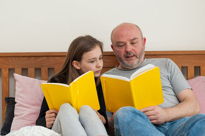 Young school age girl sitting together with her father and reading yellow textbook. solving homework