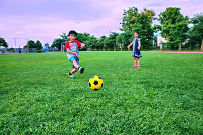 Full length of boy and girl playing soccer with ball on grass against sky