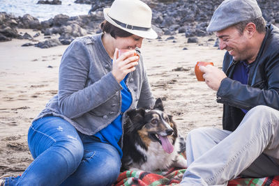 Happy couple having drinks while sitting with dog at beach