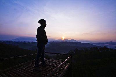Man standing by railing against sky during sunset
