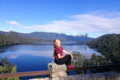 Young woman sitting on rock by lake against sky