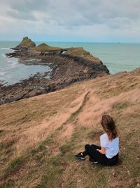 Woman sitting on grass by sea against sky