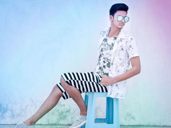 Full length of young man wearing sunglasses sitting on stool against wall