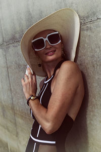 Portrait of a woman in a swimsuit, hat and sunglasses in summer on the riverbank by a concrete wall