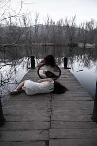 Woman lying while friend holding mirror on pier over lake