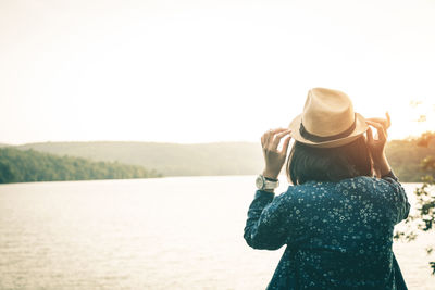 Rear view of woman wearing hat at lakeshore against clear sky during sunset