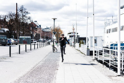 Portrait of man jumping on footpath in city against cloudy sky