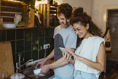 Smiling man doing dishes while looking at smart phone held by girlfriend in kitchen