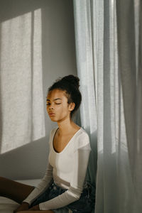 Young woman with eyes closed sitting at home against curtain
