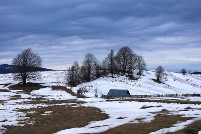 Old wooden house in the distance on a field covered in snow and dark storm clouds in the sky