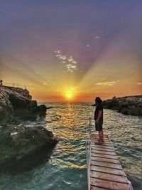 Man standing on rock by sea against sky during sunset