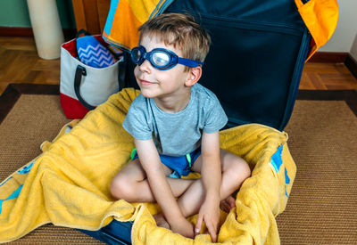 Boy wearing swimming goggles while sitting in suitcase at home