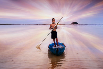 Boy rowing boat in sea against sky during sunset
