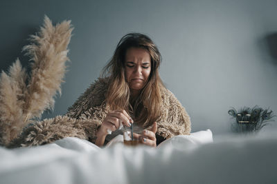 Young woman with herbal tea sitting on bed against wall