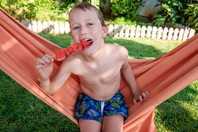Portrait of shirtless boy eating watermelon while sitting on hammock outdoors